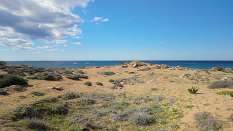 Una-Amplia-Vista-Del-Terreno-Arenoso-En-Las-Tumbas-De-Los-Reyes-En-Pafos,-Chipre,-Con-El-Mar-Al-Fondo-Y-Un-Cielo-Parcialmente-Nublado.