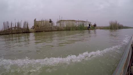 Un-Barco-Que-Navega-A-Toda-Velocidad-Por-Las-Orillas-De-Las-Marismas-En-El-Sur-De-Irak-Y-Muestra-Vistas-De-Las-Casas-Locales,-Los-Agricultores-Y-Los-Potreros-De-Búfalos-De-Agua-Hechos-Con-Juncos-Tejidos.