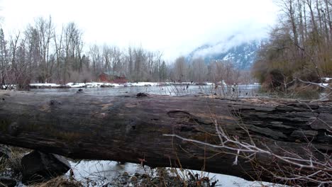 Cascade-Mountain-Pass-to-Leavenworth,-Washington---A-beautiful-Bavarian-styled-alpine-village-covered-in-snow---Fallen-log-and-creek-streaming-by---Cabin-in-the-distance