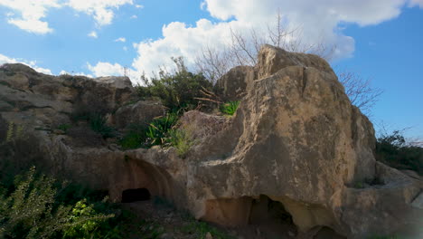 Una-Gran-Formación-Rocosa-Con-Vegetación-En-Las-Tumbas-De-Los-Reyes-En-Pafos,-Chipre,-Bajo-Un-Cielo-Parcialmente-Nublado.