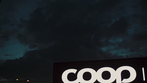 Inclínese-Hacia-Abajo-Desde-El-Cielo-Nublado-Hasta-El-Logotipo-De-La-Marca-Coop-Y-La-Entrada-Al-Hipermercado-Por-La-Noche.