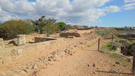 Antiguas-Ruinas-De-Piedra-En-Las-Tumbas-De-Los-Reyes-En-Pafos,-Chipre,-Con-Vegetación-Dispersa-Y-Un-Cielo-Parcialmente-Nublado