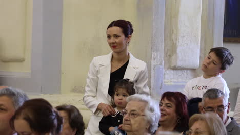 Gente-Viendo-Una-Celebración-De-Servicio-Religioso,-Un-Bebé-Comiendo-Y-Un-Niño-Pequeño-Estirándose-Durante-Una-Ceremonia