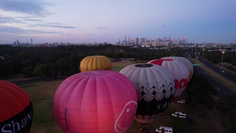 Drone-rising-over-hot-air-balloons-as-they-take-off,-revealing-Melbourne-city-in-the-backdrop-at-sunrise