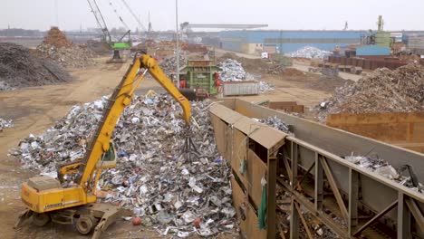 Orange-excavator-sorting-metal-scrap-at-industrial-recycling-yard,-cloudy-day