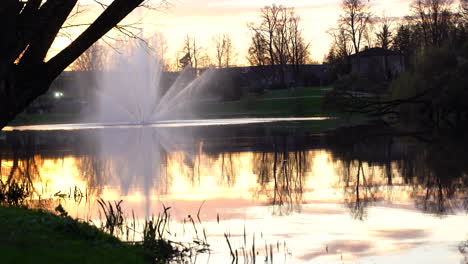 Abendlicher-Blick-Auf-Den-See-Mit-Springbrunnen-Und-Baumreflexionen-Bei-Goldenem-Sonnenuntergang