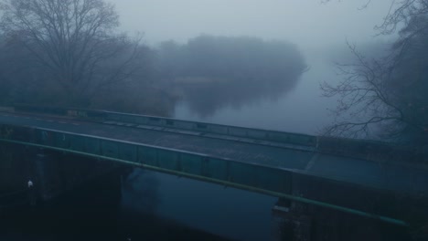 Toma-Lateral-De-Un-Coche-Cruzando-El-Puente-Con-Un-Manto-De-Niebla-Durante-Las-Primeras-Horas-De-La-Mañana.