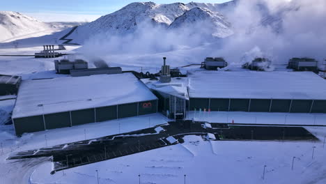 Snow-covered-geothermal-power-plant-in-Iceland,-entrance-visible,-surrounded-by-mountains,-aerial-view