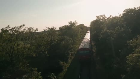 Vista-Aérea-Dinámica-Desde-Un-Dron-De-Carreras-FPV-Volando-Entre-árboles-Persiguiendo-Un-Tren-De-Pasajeros-De-Color-Rojo-Que-Cruza-El-Río-Morava-En-Un-Viejo-Puente-De-Hierro.