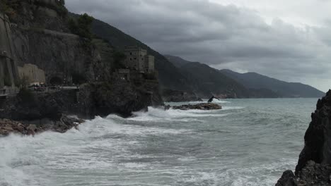 Toma-Aérea-De-Un-Dron-De-La-Ola-De-Cinque-Terre-Rompiendo-En-Una-Roca-En-La-Playa-Zoom-En-El-Castillo-De-Un-Pueblo-Italiano-En-El-Acantilado