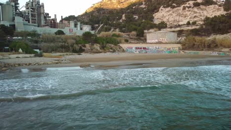 Costa-garraf-beach-with-waves,-graffiti,-and-a-factory-in-the-background,-aerial-view