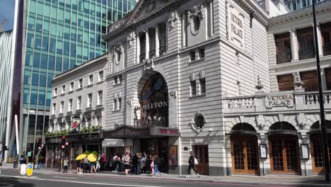 Teatro-Victoria-Palace-En-Londres-Durante-El-Día,-Con-Gente-En-Las-Aceras-Y-Tráfico-Pasando,-Mostrando-El-Concepto-De-Ajetreo-Urbano-Y-Monumentos-Culturales