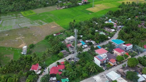 Orbiting-aerial-overview-of-radio-communication-tower-in-tropical-island-village-with-small-neighborhood-and-rice-fields-in-Catanduanes,-Philippines