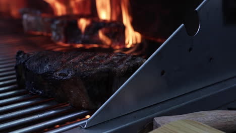 Flames-rising-on-barbeque-behind-well-done-steak-in-foreground,-close-up-shallow-focus-slow-motion