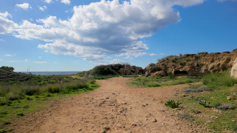 A-dirt-pathway-leading-through-the-archaeological-site-of-the-Tombs-of-the-Kings-in-Pafos,-with-ancient-ruins-and-greenery-on-either-side