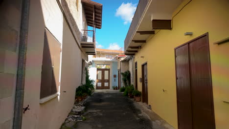 Eine-Schmale-Gasse-In-Einem-Traditionellen-Dorf-Mit-Pastellfarbenen-Wänden,-Topfpflanzen-Und-Einem-Strahlend-Blauen-Himmel