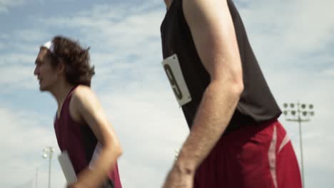 low-angle-tracking-shot-of-tired-runners-on-a-hot-sunny-day-sprinting,-wearing-maroon-uniforms