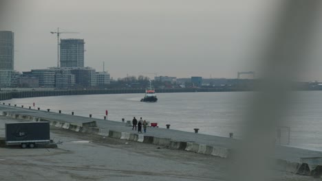 Un-Velero-En-El-Puerto-De-Amberes-Con-Peatones-Caminando-Y-Un-Remolque-En-El-Estacionamiento-Y-Grúas-Al-Fondo-Junto-A-Edificios-De-Paneles-Con-Apartamentos