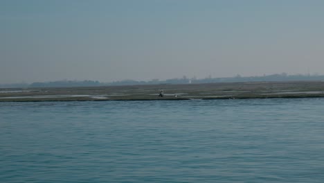 Ruhiger-Blick-Von-Einem-Boot-In-Venedig,-Der-Das-Ruhige-Wasser-Und-Eine-Entfernte,-Tief-Liegende-Insel-Unter-Einem-Klaren-Blauen-Himmel-Einfängt