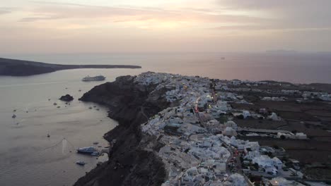 DRONE-SHOT-OF-SANTORINI-GREECE-CLIFFSIDE-AND-WHITE-HOUSES-AT-SUNSET-BOATS-MARINA-4K