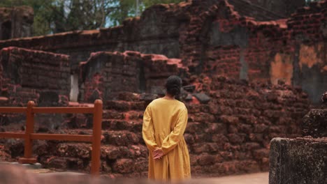 Woman-in-yellow-dress-exploring-ancient-ruins,-standing-thoughtfully-with-hands-behind-her-back