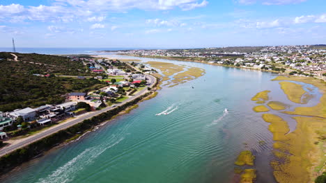 Motorized-watercraft-speeding-on-scenic-Goukou-estuarine-river-in-Still-Bay-Western-Cape-South-Africa,-aerial-riser-shot-at-sunrise