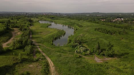 Aerial-view-of-a-lake-surrounded-by-lush-greenery,-with-paths-and-distant-buildings