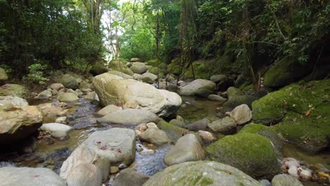 A-low-drone-shot-passing-over-large-boulders-and-rocks-following-a-river-of-fresh-water-surrounded-by-trees-in-a-rainforest,-Santa-Marta,-Colombia