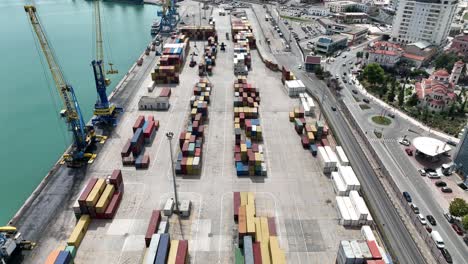 Aerial-view-of-a-shipping-cargo-dock-with-large-containers-near-a-busy-street,-reveal-wide-shot