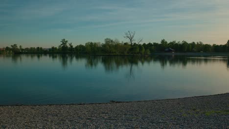 Ruhiger-See-Mit-Baumreflexionen,-Kieselstrand-Und-Klarem-Himmel-Am-Jarun-See,-Zagreb,-Kroatien