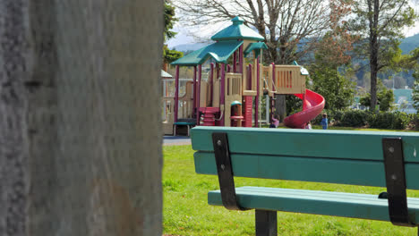 Mutter-Geht-Auf-Kleinkinder-Zu,-Die-Auf-Einem-Spielplatz-Im-Park-Spielen,-Bank-Im-Vordergrund