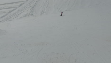 Drone-view-of-snowboarder-walking-on-the-pistes-on-the-snowy-slopes-of-Mount-Hermon,-Golan-Heights,-Israel,-flying-backward