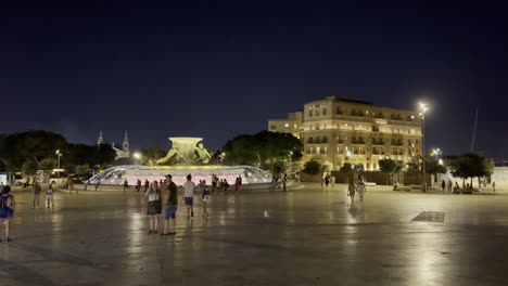 Plaza-De-La-Fuente-Tritón-Frente-A-La-Puerta-De-La-Ciudad-Por-La-Noche-Mientras-Los-Turistas-Caminan