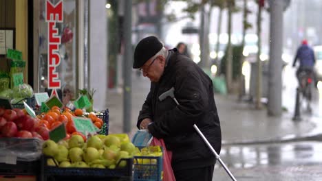 Old-man-holds-cane-and-grabs-fruits-from-roadside-stand-with-plastic-bag