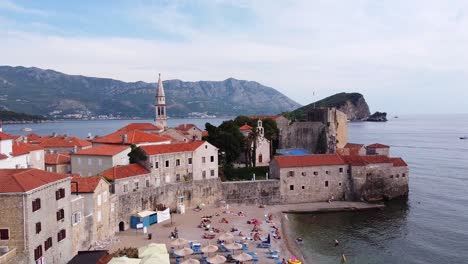 Budva,-Montenegro---Aerial-Drone-View-of-the-Boulevard,-Beach,-Sunbeds,-Tourists,-City-Wall,-Ancient-Town,-Island,-Boats,-Mountains-and-Island