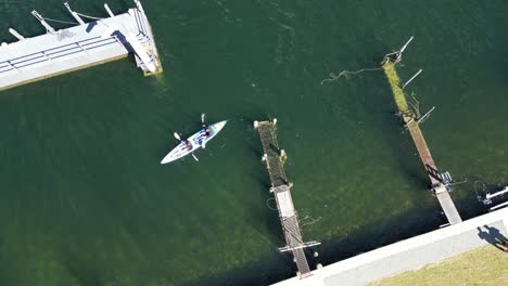 Dos-Personas-Reman-En-Kayak-En-Un-Río-Sereno-Cerca-De-Un-Muelle-En-Nueva-Zelanda,-Cerca-De-La-Ciudad-De-Te-Anau.