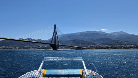 POV-Sailing-at-Greek-blue-sea-slow-motion-motorboat-front-reveals-Rio-Bridge,-mountain-skyline-background-at-shiny-daylight