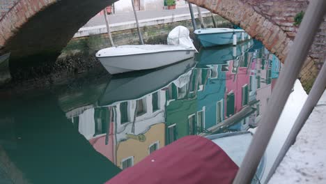 Picturesque-view-of-boats-docked-under-a-bridge-in-Burano-Island,-Venice,-with-colorful-buildings-reflecting-on-the-calm-water