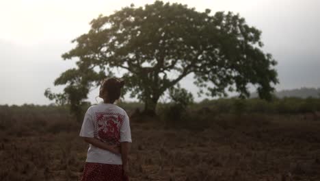 Young-woman-stands-in-field,-gazing-at-tree-during-sunset