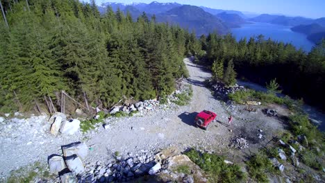 reverse-revealing-aerial-of-a-Jeep-parked-on-a-loggin-road-surrounded-by-trees-and-lakes-in-the-background