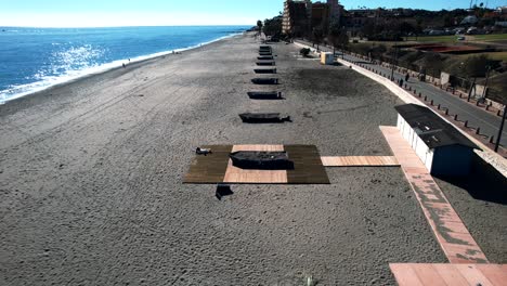 Aerial-view-over-the-beach-of-Fuengirola,-Spain-with-boats-laying-around-the-beach-and-a-higher-angle-view-of-the-city-highway-road