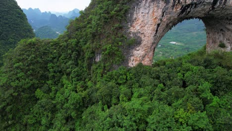 Aerial-view-of-Moon-Hill-arch-in-lush-Yangshuo-with-a-person-exploring-below,-China