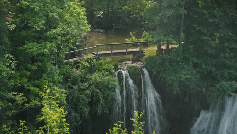 Beautiful-dual-waterfall-with-a-wooden-footbridge-and-lush-greenery-in-Rastoke,-Croatia