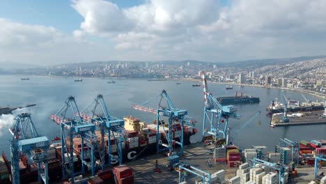 Aerial-orbit-of-a-cargo-ship-docked-near-cranes-and-containers,-Valparaiso-Sea-Port-and-city-in-background,-Chile