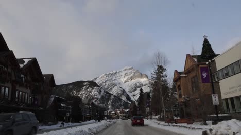 Cruzando-La-Avenida-Banff-Con-La-Montaña-En-Cascada-En-La-Distancia,-Paisaje-Invernal-Pov-Del-Conductor
