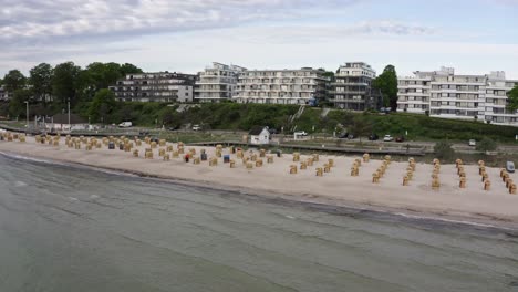 Aerial-pan-view-from-a-drone-of-Scharbeutz-Beach,-Germany,-a-resort-town-in-Schleswig-Holstein-on-the-Baltic-coast,-with-chairs-overlooked-by-high-rise-waterfront-buildings