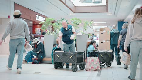 Ein-Weißer-Mann-In-Einer-Blauen-Weste-Mit-Einem-Einkaufswagen-Hält-An,-Um-Seine-Artikel-Auf-Dem-Jährlichen-Buchmarkt-Des-Kinderkrankenhauses-Im-Einkaufszentrum-Zu-Zählen