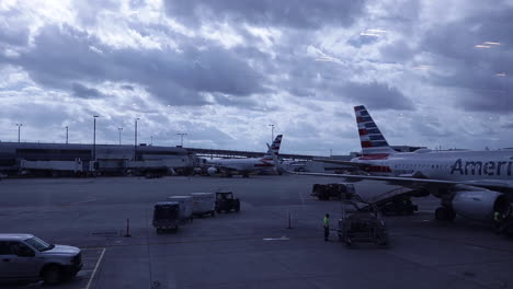 Airport-luggage-cart-driving-to-airplane-parked-at-gate-on-cloudy-grey-day,-static