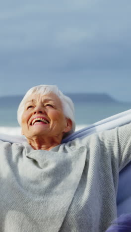 Senior-woman-standing-with-hands-raised-on-beach