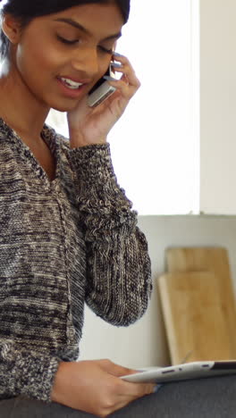 Woman-talking-on-mobile-phone-while-using-digital-tablet-in-kitchen